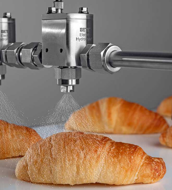 Stainless steel BETE spray nozzles spray coating on croissants moving along on a white conveyor.