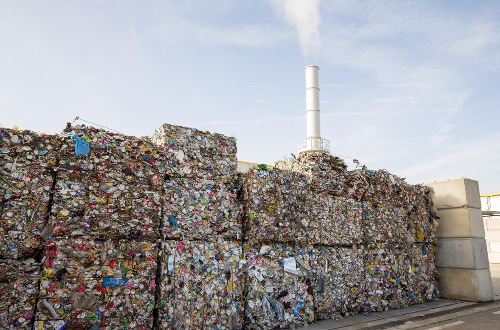 Large bales of crushed cans stacked outside of a waste-to-energy plant with a chimney flue in the background emitting steam or smoke into the air.